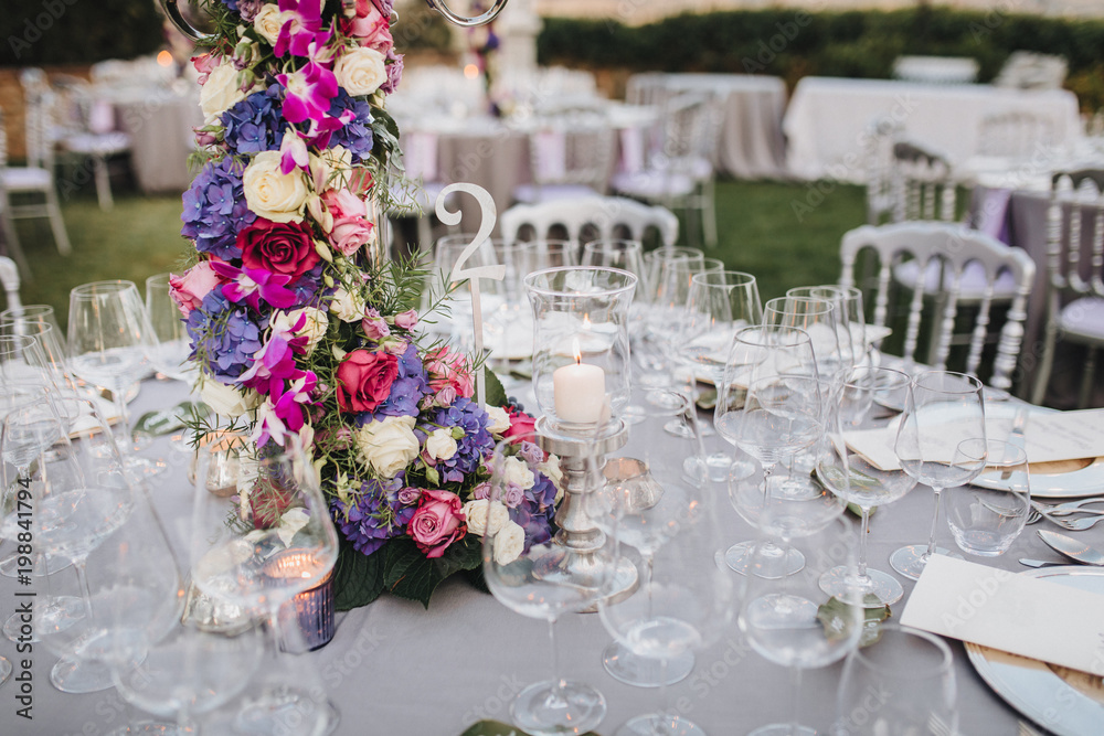 the flower arrangement stands on the table in the wedding banquet area on a green glade, the table is decorated with candles, there are plates, glasses, cutlery the flower arrangement stands on the table in the wedding banquet area on a green glade, the table is decorated with candles, there are plates, glasses, cutlery