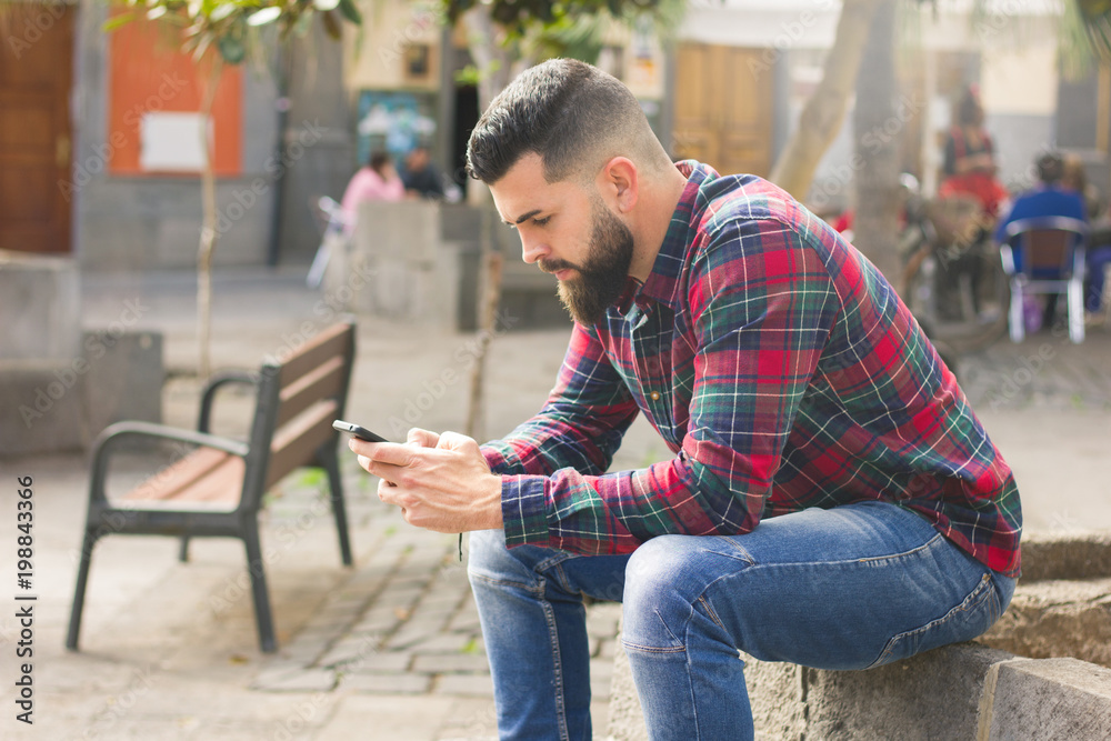Handsome man with full beard and red checkers flannel shirt holding smart phone in Las Palmas old town square, Spain. Young hipster using mobile device sitting on sunny day in the city Handsome man with full beard and red checkers flannel shirt holding smart phone in Las Palmas old town square, Spain. Young hipster using mobile device sitting on sunny day in the city