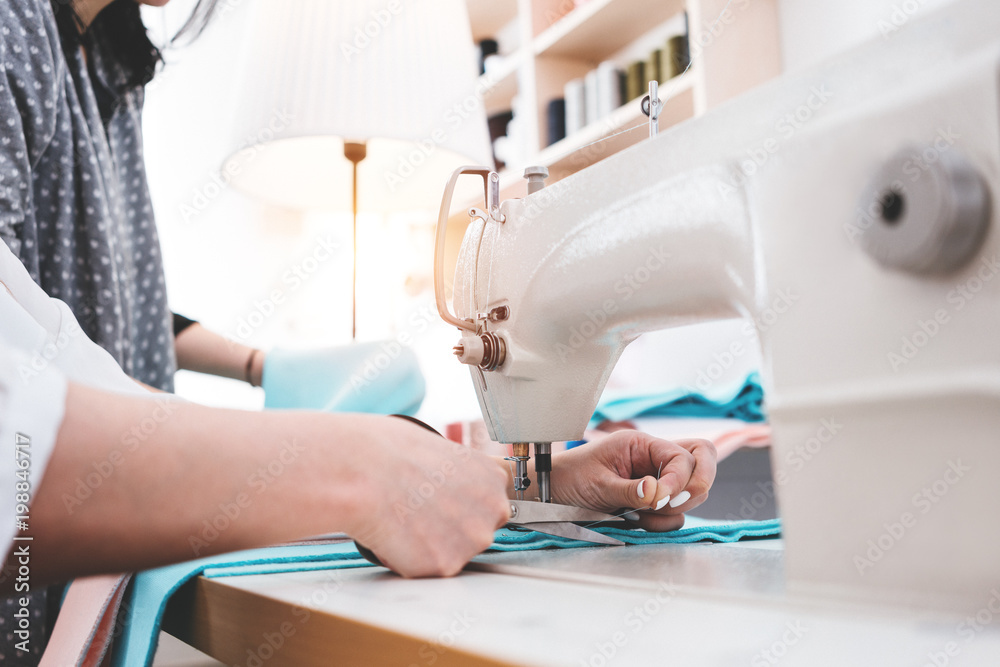 Creative dressmaker woman working with cloth on sewing machine. Two seamstress in design bureau making new clothing concept. Small business. Flare effect. Blurred background