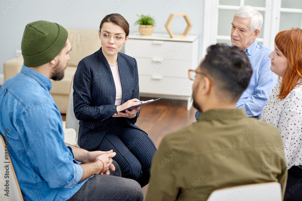 Fototapeta premium Group therapy session at full speed: confident young psychologist discussing problem with patient and taking notes on clipboard while other participants listening to them