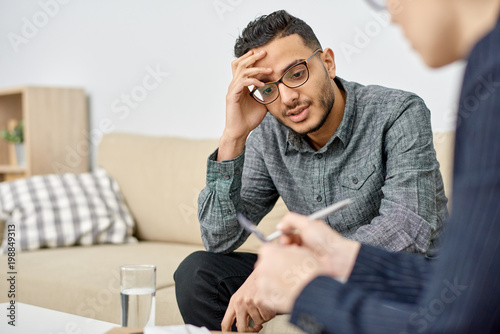 Fototapeta Naklejka Na Ścianę i Meble -  Close-up shot of young psychologist taking notes on clipboard while consulting depressed mixed-race patient during therapy session at cozy office