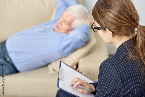 Senior patient lying on cozy sofa with hands on nape and sharing his problems with highly professional psychologist while having therapy session