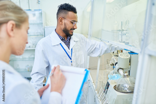 Positive content confident young Arabian scientist in lab coat adjusting laboratory equipment and reporting medical assistant about results