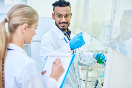 Cheerful skilled handsome young Arabian male chemist with beard holding laboratory glassware and explaining experiment to young colleague