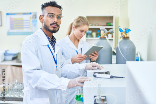 Serious handsome young Arabian scientist in lab coat choosing program for machine while working in laboratory, his female assistant using tablet behind him