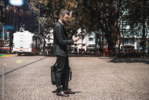 Young handsome bearded businessman in a formal suit and glasses is standing on a pavement stone outdoors, calling taxi via an app on his smartphone; with copy space place for advertising or your text