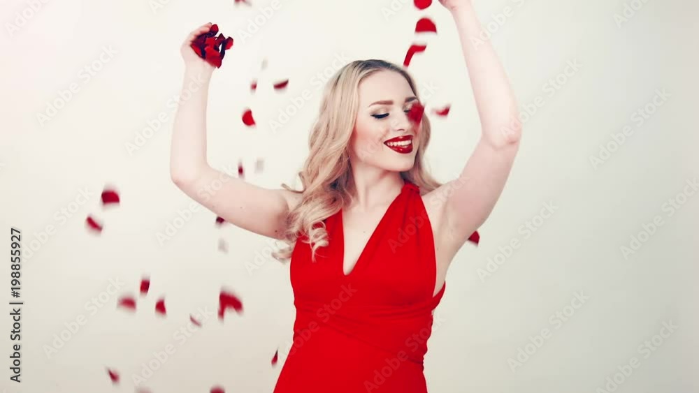 Fashionable blonde beauty in red dress among falling rose petals. Petals red roses fall on girl with festive red dress. In studio. Beautiful makeup and red lipstick. Sincerely smiles and rejoices. Fashionable blonde beauty in red dress among falling rose petals. Petals red roses fall on girl with festive red dress. In studio. Beautiful makeup and red lipstick. Sincerely smiles and rejoices.