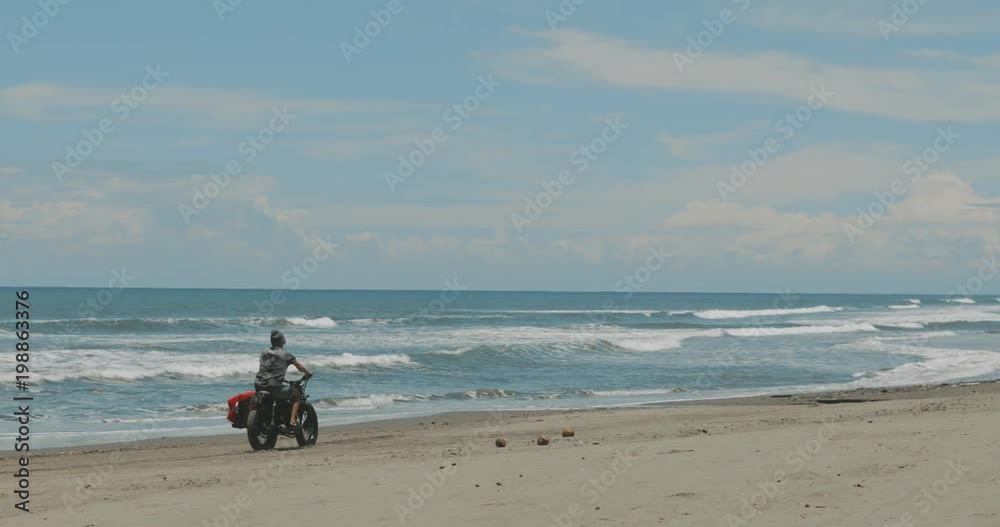 Handsome man biker surfer driving his black motorcycle cafe racer with red surfboard shortboard on beach along ocean at sunny day. 4k video shooting by handheld gimbal Handsome man biker surfer driving his black motorcycle cafe racer with red surfboard shortboard on beach along ocean at sunny day. 4k video shooting by handheld gimbal