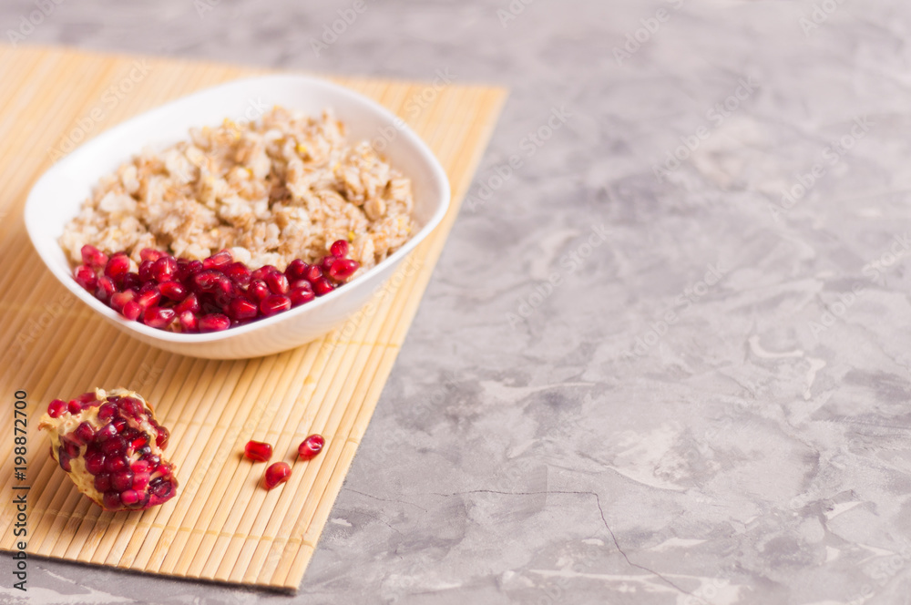 Cooked yummy warm oatmeal and heap of ripe fresh pomegranate seeds in white ceramic bowl near piece of pomegranate on clean bamboo rug on worn gray scratched concrete with copy space Cooked yummy warm oatmeal and heap of ripe fresh pomegranate seeds in white ceramic bowl near piece of pomegranate on clean bamboo rug on worn gray scratched concrete with copy space