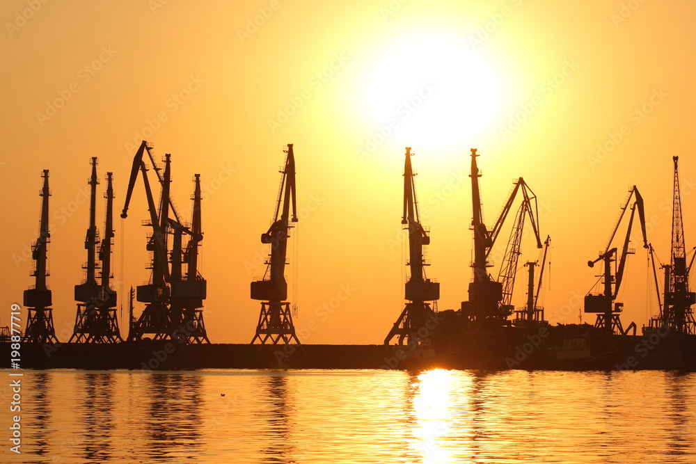 Seaport at sunset, cranes in the port on the horizon, orange sky against the sea horizon, landscape with high-rise cranes, construction of a breakwater in the sea