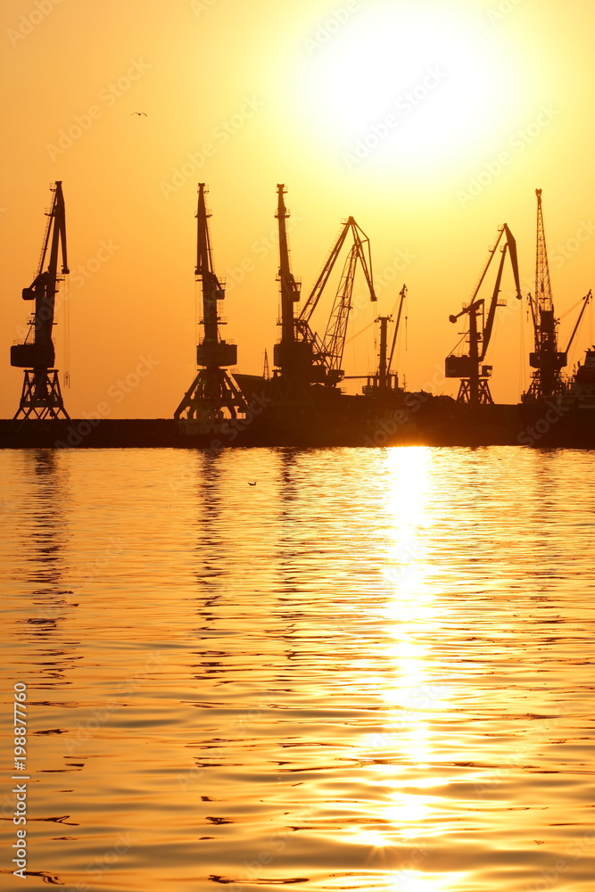 Seaport at sunset, cranes in the port on the horizon, orange sky against the sea horizon, landscape with high-rise cranes, construction of a breakwater in the sea
