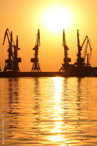 Seaport at sunset, cranes in the port on the horizon, orange sky against the sea horizon, landscape with high-rise cranes, construction of a breakwater in the sea