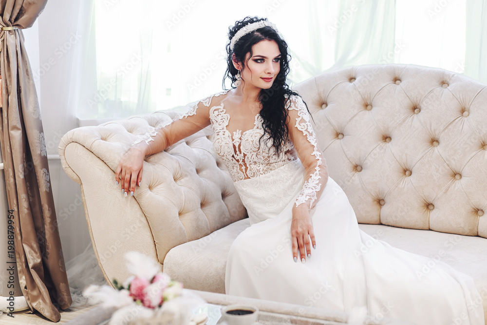 the bride in a lace dress sits on the couch in the morning