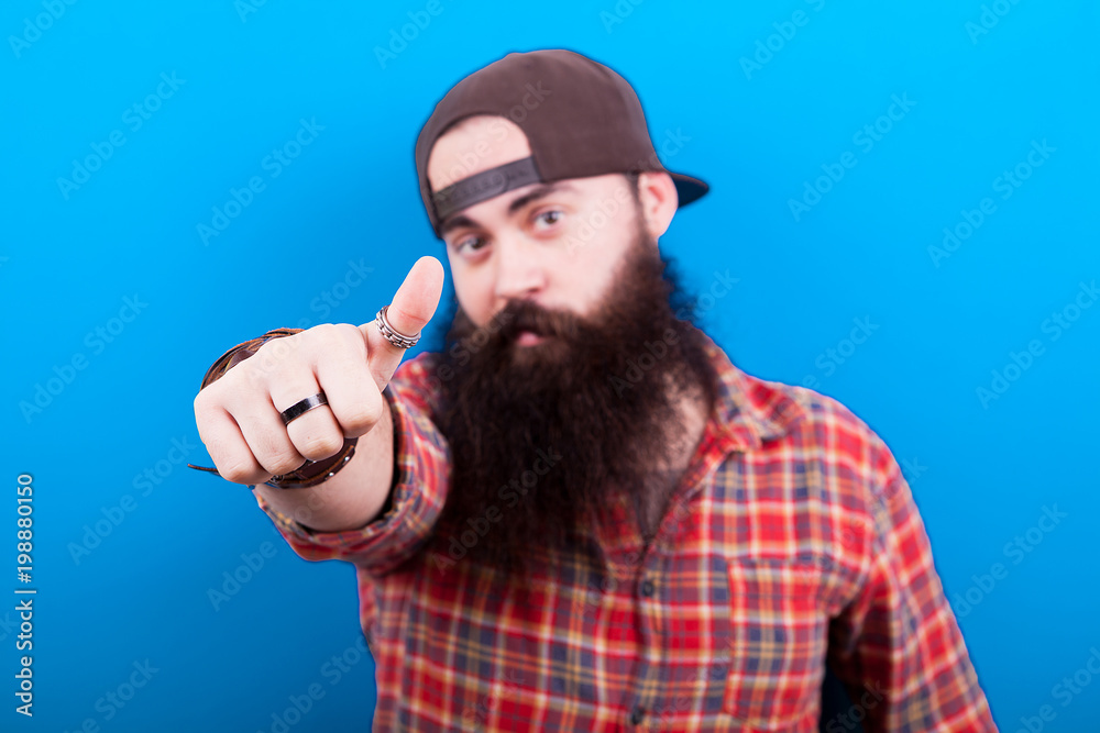 Naklejka premium Long bearded man showing thumbs up to the camera and only the hand is in focus. Blue background in studio photo