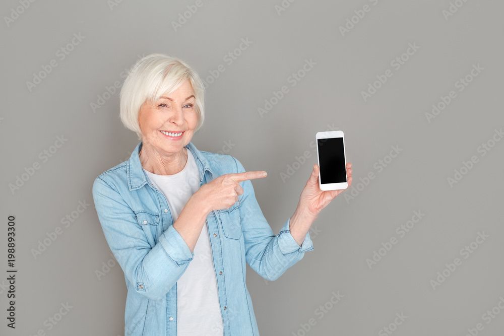 Senior woman in jeans jacket studio isolated on grey wall pointing at smartphone screen Senior woman in jeans jacket studio isolated on grey wall pointing at smartphone screen
