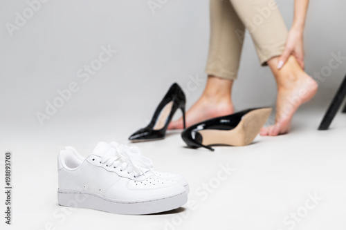 Tired woman touching her ankle, suffering from leg pain because of uncomfortable shoes, feet pain wear high heel shoes after walk, focus on comfortable sneakers