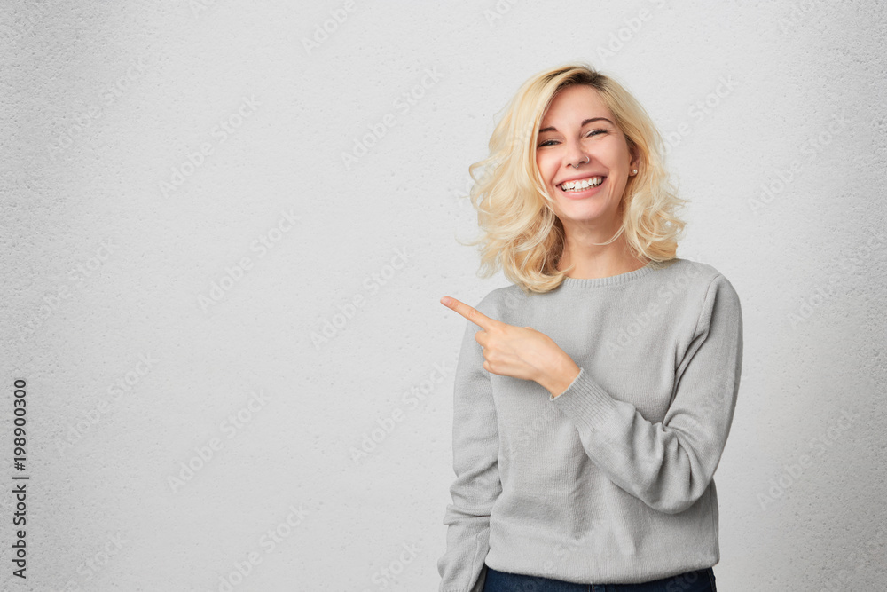 Happy excited female with blonde hair indicates with fore finger aside, demonstrates something at blank copy space for your advertisment or hearder. Positive expression. Isolated Happy excited female with blonde hair indicates with fore finger aside, demonstrates something at blank copy space for your advertisment or hearder. Positive expression. Isolated