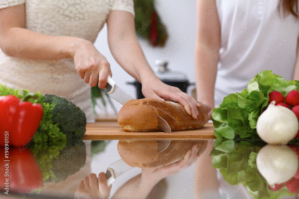 Closeup of human hands cooking in kitchen. Mother and daughter or two female cutting bread at the table full of vegetables and green salad. Healthy meal, vegetarian food and lifestyle concepts Closeup of human hands cooking in kitchen. Mother and daughter or two female cutting bread at the table full of vegetables and green salad. Healthy meal, vegetarian food and lifestyle concepts