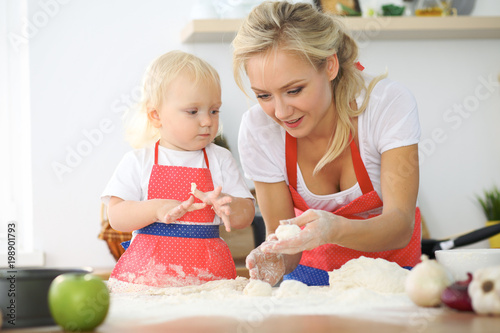 Little girl and her blonde mom in red aprons playing and laughing while kneading the dough in the kitchen. Homemade pastry for bread, pizza or bake cookies