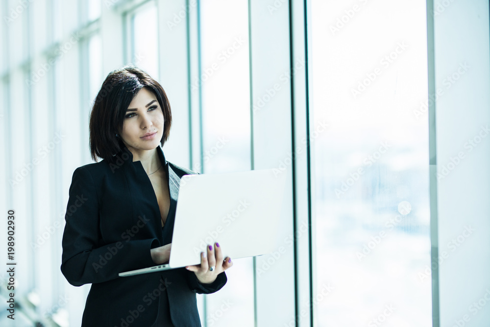 Portrait of young business woman working holding laptop standing against panoramic window with city view Portrait of young business woman working holding laptop standing against panoramic window with city view