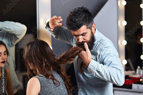 Professional hairdresser working with fashion brunette model with long curly hair at luxury salon. Barbershop. Barber scissors. Womens haircut. Bearded man, lush beard. Barber shop service concept.