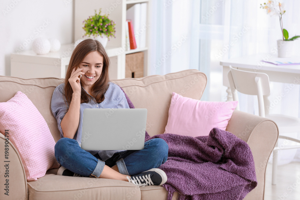 Young woman talking on mobile phone while working with laptop at home