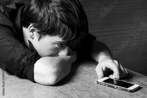 Worried man laying on a table, looking to his smart phone. Bad news and fear. Converted to black and white. Grain added.
