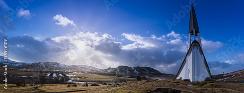 Panorama of the Mosfellsbaer church in Iceland at sunset with a cross, graves, green grass, mountains, clouds, and blue sky