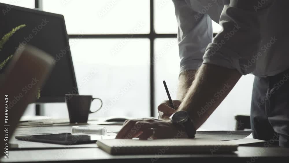 Male hands of an architect against the background of a large window. Man makes notes, writes with a pencil, leaning on the table: close-up. Man works in loft office with papers, drawings and documents Male hands of an architect against the background of a large window. Man makes notes, writes with a pencil, leaning on the table: close-up. Man works in loft office with papers, drawings and documents