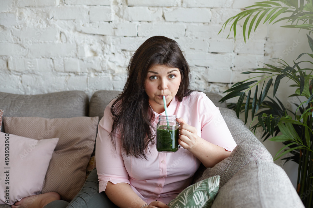 Portrait of attractive plus size young brunette woman with curvy body hoping to loose extra pounds, drinking detox fresh juice every morning, seeping green drink using straw, sitting in living room Portrait of attractive plus size young brunette woman with curvy body hoping to loose extra pounds, drinking detox fresh juice every morning, seeping green drink using straw, sitting in living room