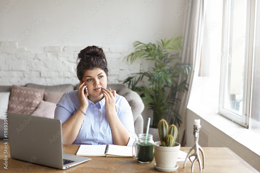 Horizontal shot of concentrated chubby young woman with hair bun multitasking at home office, sitting at desk in front of open laptop computer, making notes in diary and having phone conversation Horizontal shot of concentrated chubby young woman with hair bun multitasking at home office, sitting at desk in front of open laptop computer, making notes in diary and having phone conversation
