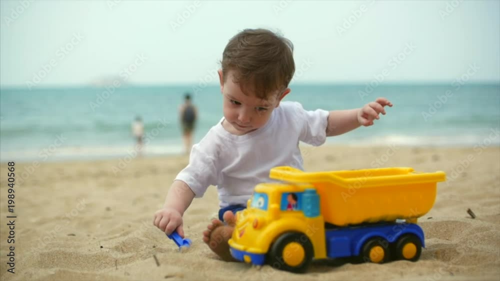 Two-year old beautiful baby toddler boy riding a toy cargo car on summer beach closeup. Slow motion Two-year old beautiful baby toddler boy riding a toy cargo car on summer beach closeup. Slow motion