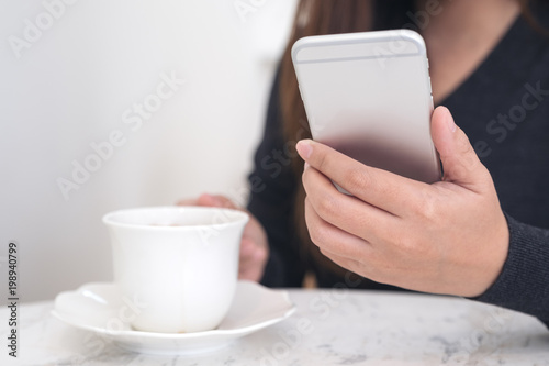 Closeup image of a woman holding and using smartphone while drinking coffee in cafe
