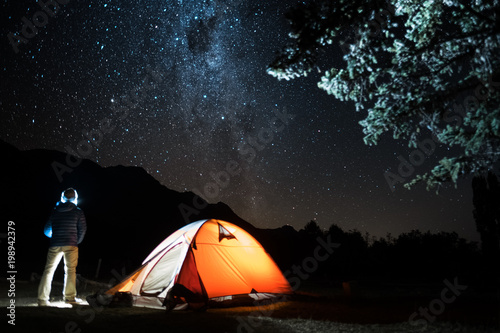 Hiker stands by the tent and enjoys night starry sky