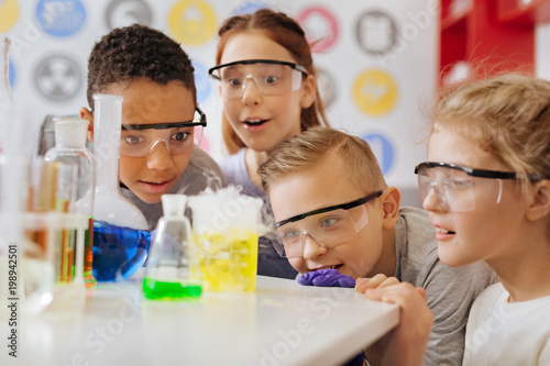 Miracles of chemistry. A group of cheerful teenage students squatting near the table and observing the chemical reaction going on in the flask with excited and surprised looks on their faces