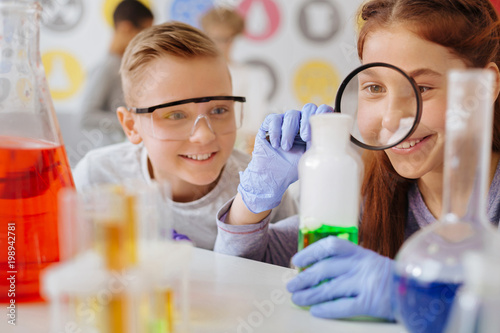 Thorough examination. Upbeat teenage girl examining a green substance in the flask with the magnifying glass while her classmate watching her do so with a smile