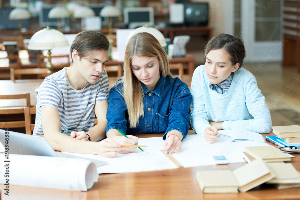Group of pensive concentrated architecture students discussing blueprint while sitting at table in library Group of pensive concentrated architecture students discussing blueprint while sitting at table in library