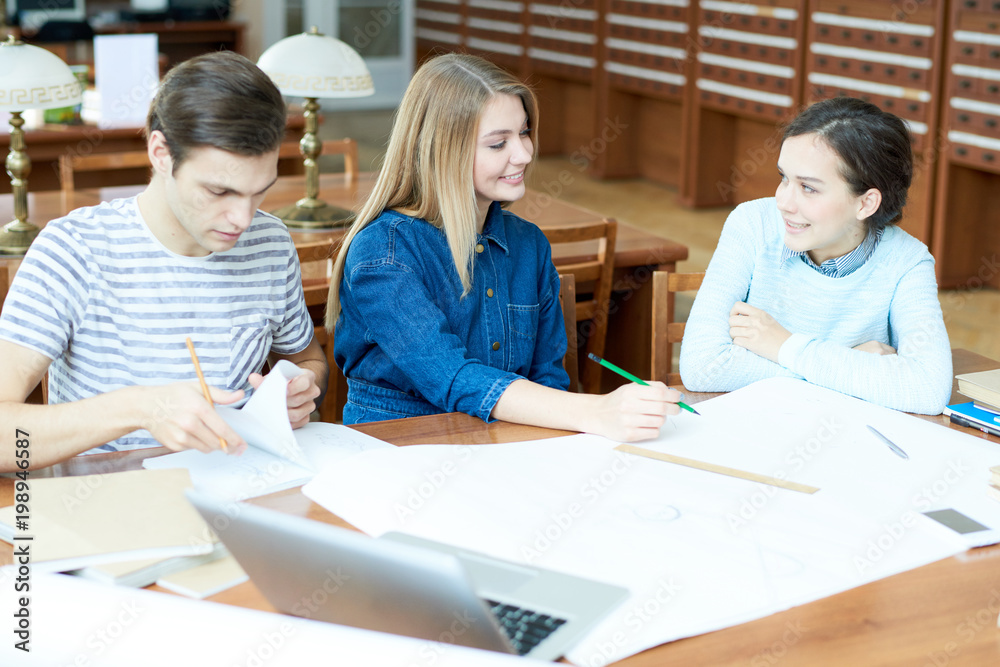Group of positive optimistic friends drawing sketch together discussing classes in library after university and sitting at table with blueprint Group of positive optimistic friends drawing sketch together discussing classes in library after university and sitting at table with blueprint