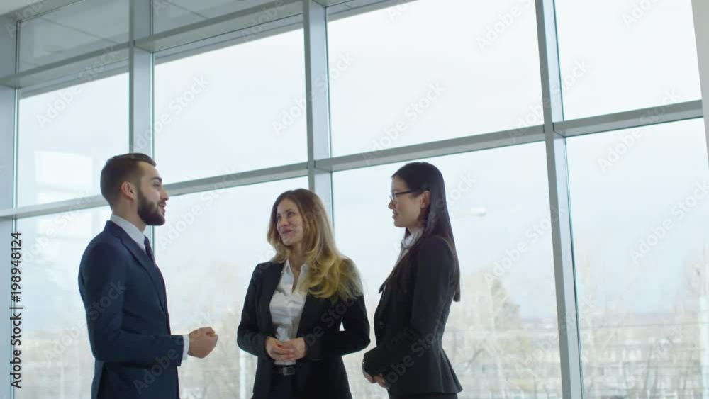 Medium shot of handsome bearded businessman in suit chatting with female colleagues in office with panoramic windows Medium shot of handsome bearded businessman in suit chatting with female colleagues in office with panoramic windows