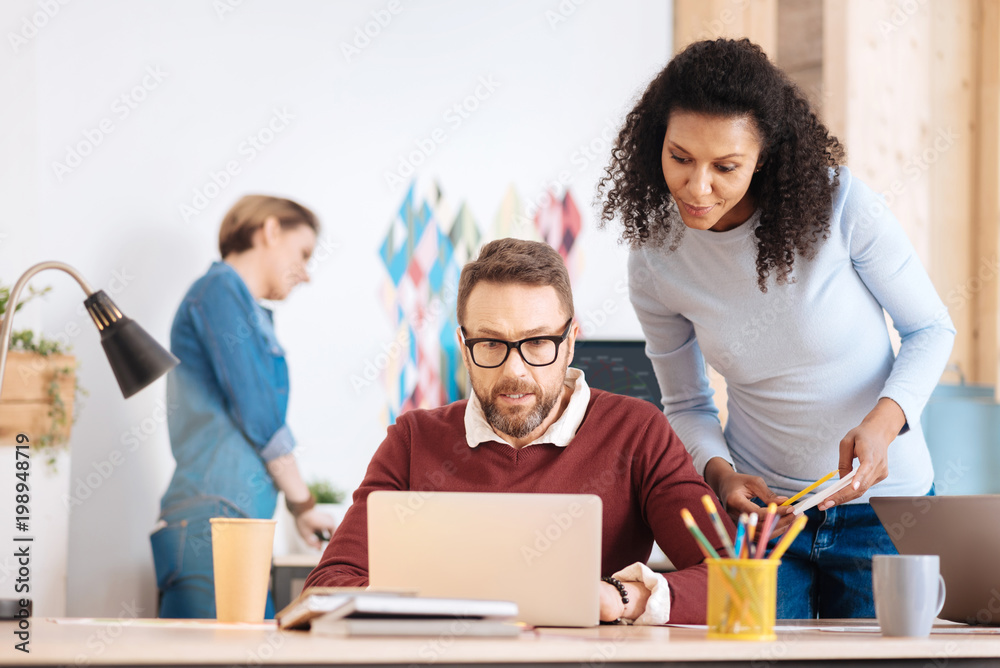 My working day. Handsome concentrated bearded man working on his laptop while sitting at the table and his colleague standing behind him My working day. Handsome concentrated bearded man working on his laptop while sitting at the table and his colleague standing behind him