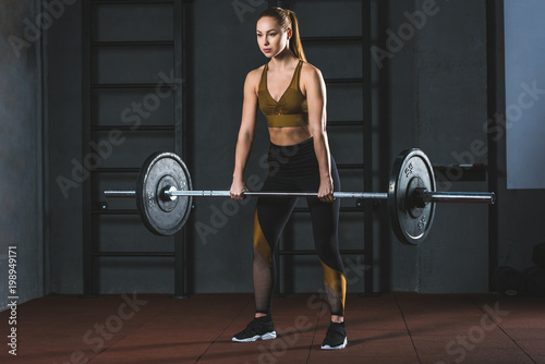 Front view of young sportswoman exercising with barbell in sports hall