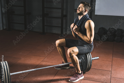 Tired sportsman with towel sitting on barbell in gym
