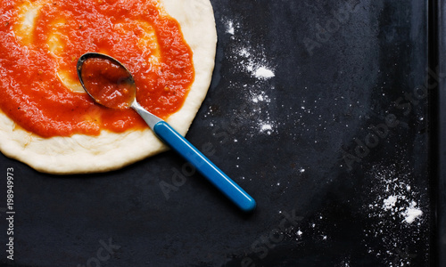 Preparation of homemade pizza. Cooking process. Smearing ketchup on dough. Top view on dark metallic background.