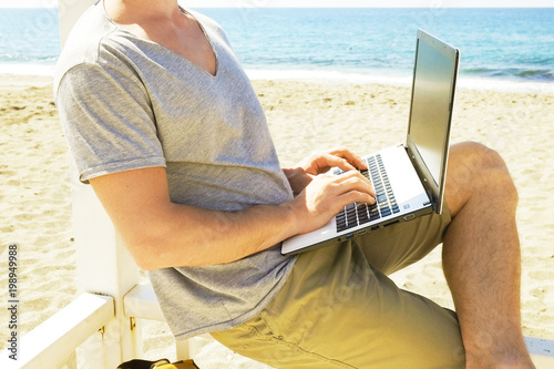Young man in grey shirt sitting at the beach, working on laptop. Blogger, writer, coder, remote worker surfing browsing researching on notebook. Wireless internet on sea shore. Background, copy space