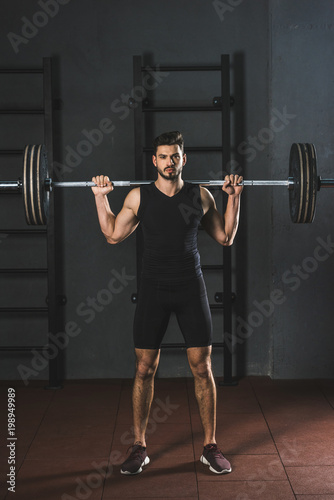 Front view of young sportsman holding barbell on shoulders in gym