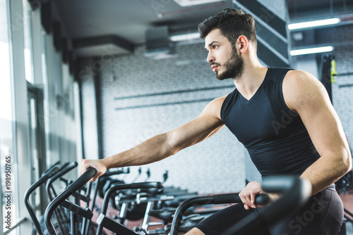Side view of young sportsman doing workout on exercise bike in sports center