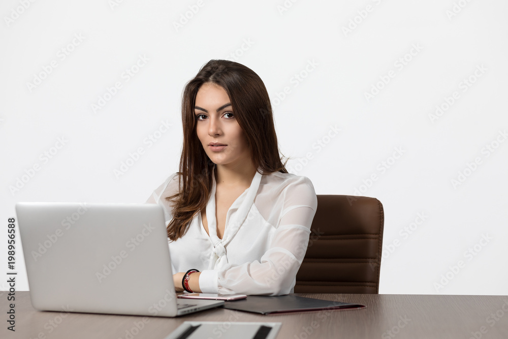 Beautiful young business woman looking at camera and smiling while working at laptop in office. Woman against white background. Beautiful young business woman looking at camera and smiling while working at laptop in office. Woman against white background.