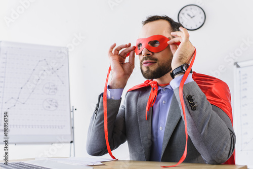 super businessman in cape looking through red mask in office