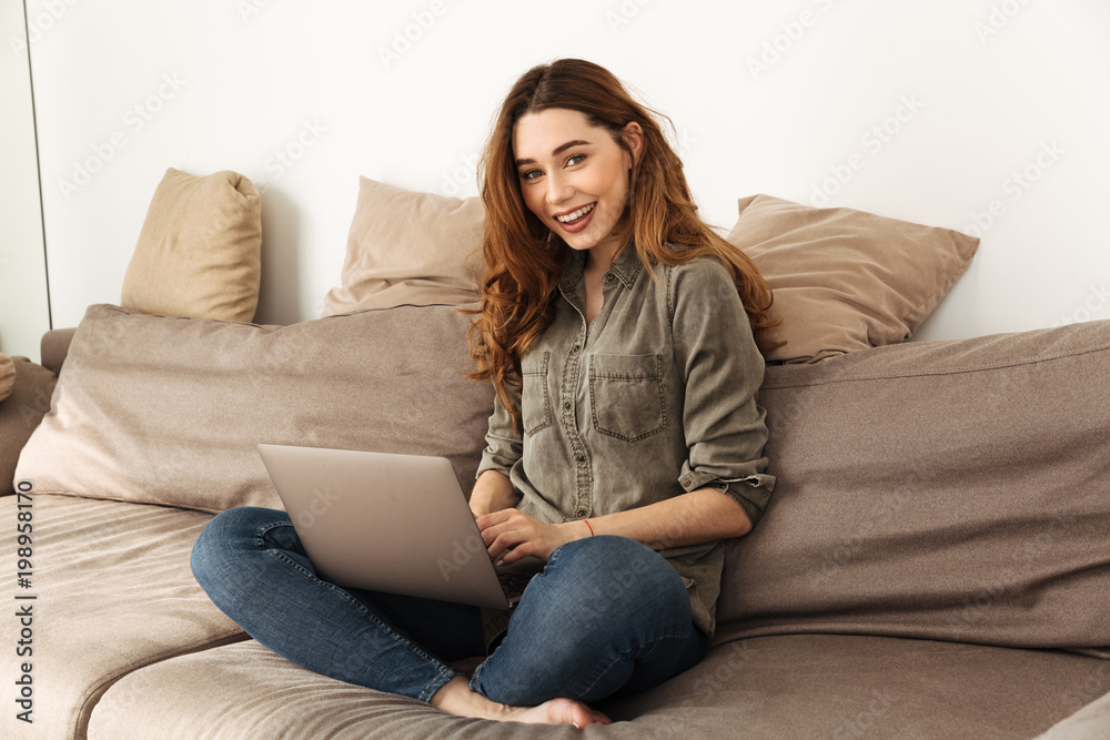 Image of relaxed affable woman with brown hair sitting on sofa in living room with legs crossed, and using laptop looking on camera Image of relaxed affable woman with brown hair sitting on sofa in living room with legs crossed, and using laptop looking on camera