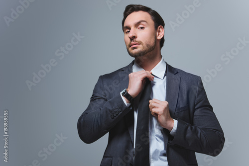 handsome businessman fixing tie and looking at camera isolated on grey
